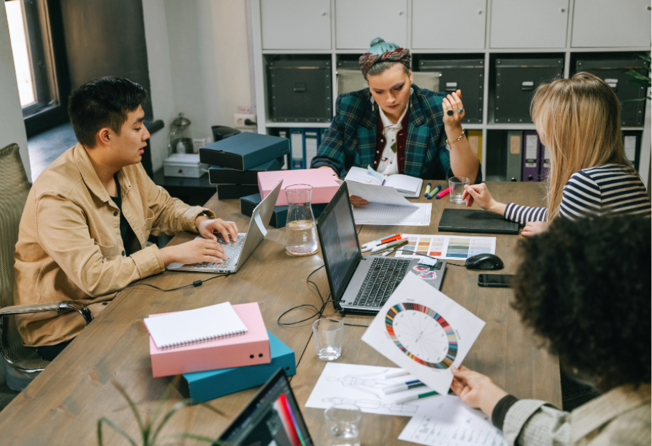 A diverse team collaborates around a shared office table, reviewing documents, charts, and project materials while using laptops, creating a productive and creative workplace atmosphere.